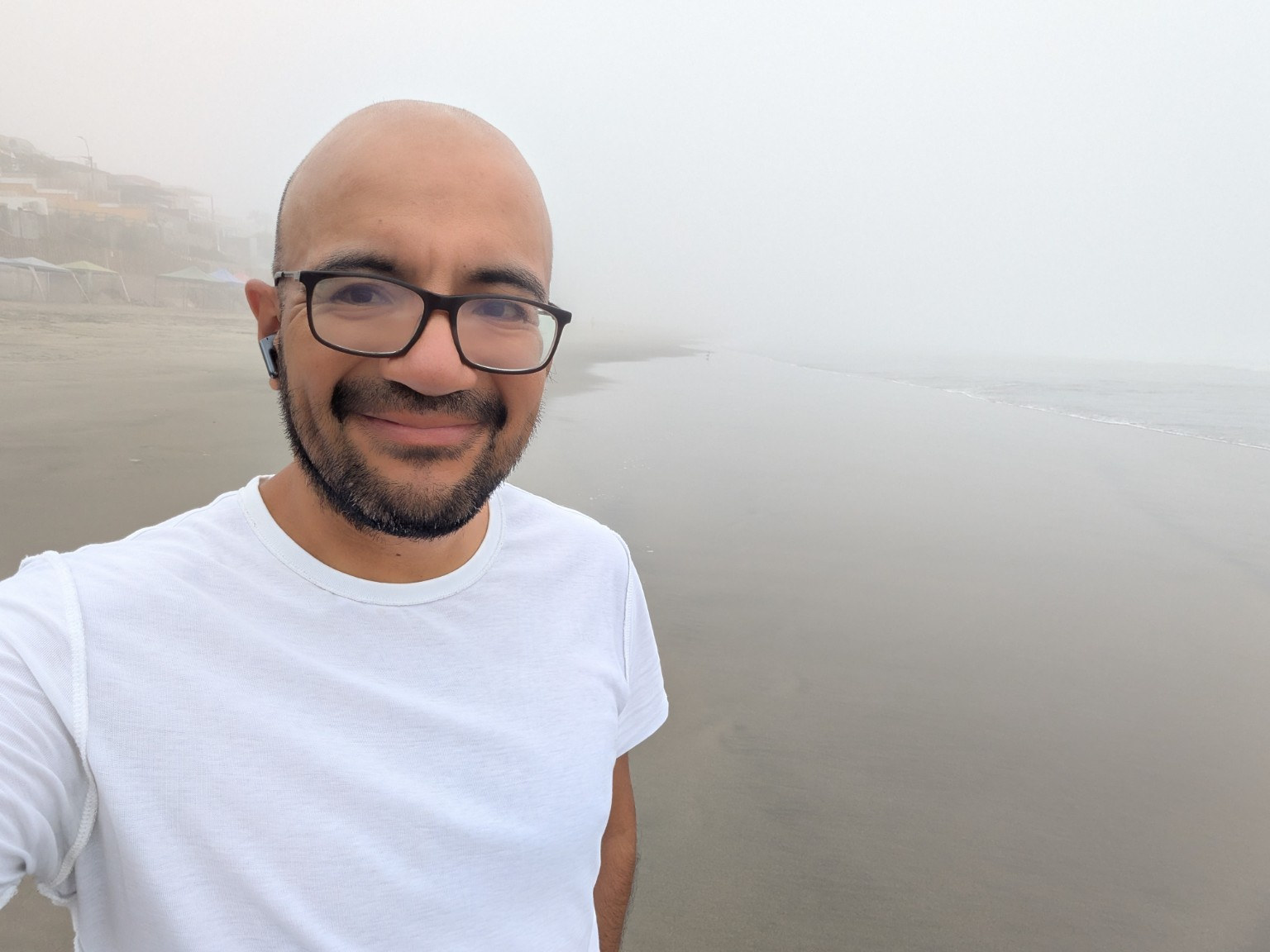 Omar Manky, smiling, wearing glasses and a white T-shirt, on a beach covered in fog. Lima, winter 2024.